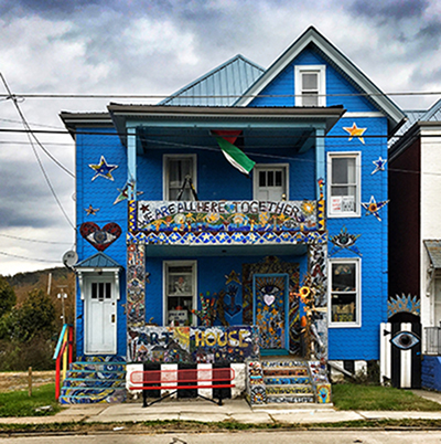 Figure 6 Image of a two-story home painted bright blue and covered in art from top to bottom. The words “WE ARE ALL HERE TOGETHER” adorn the mosaic artwork on the second-floor balcony.